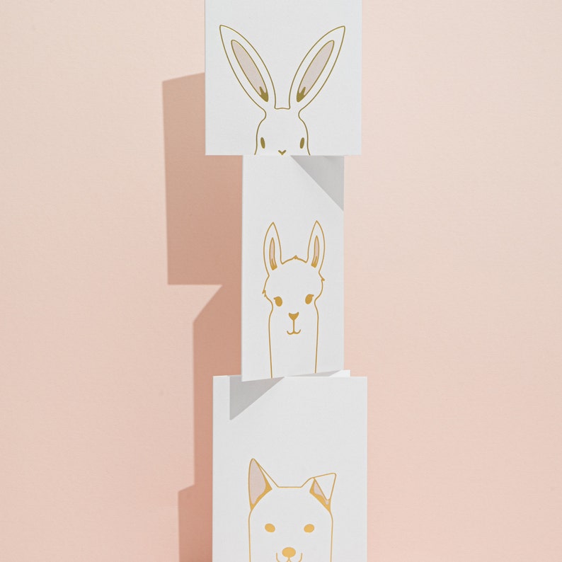 May include: Three stacked white cards with gold line drawings of animal faces: a rabbit, a llama, and a dog. The rabbit has long ears, the llama has a small smile, and the dog has pointed ears. The background is a solid light pink.