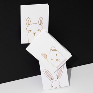 May include: White greeting cards with gold animal illustrations. The cards feature a llama, a dog, and a rabbit. The illustrations are simple line drawings with minimal detail, set against a black and white background.