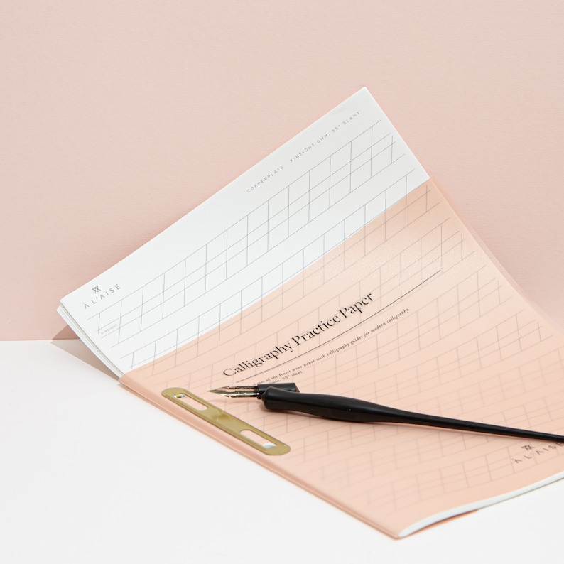 May include: Calligraphy practice paper with a black pen and a gold-colored pen holder. The paper is light pink and white with grid lines and the text "Calligraphy Practice Paper". The pen has a black handle and a metal nib. The paper is labeled "ALAISE".