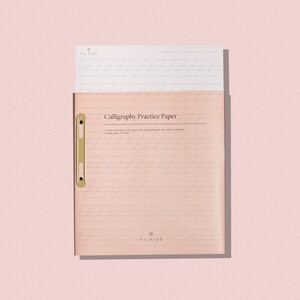 May include: A blush pink calligraphy practice paper pad with a gold-colored clasp. The pad contains 25 sheets of fine paper with calligraphy guides. The text "Calligraphy Practice Paper" is printed on the cover, along with the brand name "ALAISE".