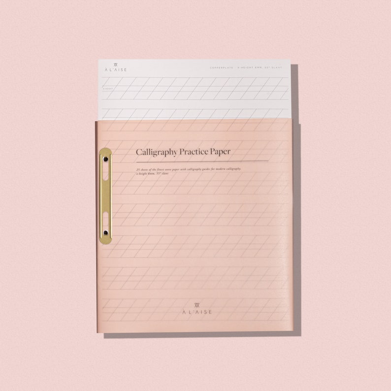 May include: A blush pink calligraphy practice paper pad with a gold-colored clasp. The pad contains 25 sheets of fine paper with calligraphy guides. The text "Calligraphy Practice Paper" is printed on the cover, along with the brand name "ALAISE".