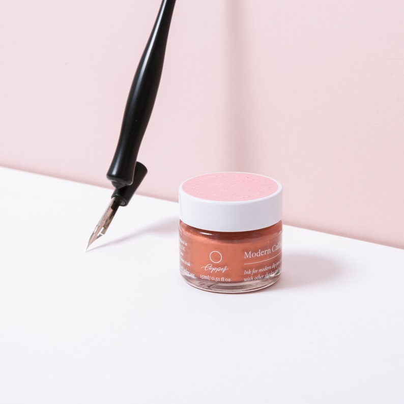 May include: A black calligraphy pen is positioned near a small glass jar of copper-coloured ink with a white lid and pink top. The jar has the text "Modern Calligraphy" and "15ml 0.51 fl oz". The background is a soft pink.