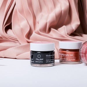 May include: Calligraphy ink jars in black, copper, and pink with white lids. A calligraphy pen is on the left. The jars are labelled "Modern Calligraphy Ink" and "Fine Gold". The background is a draped, pleated fabric in a pale pink colour.