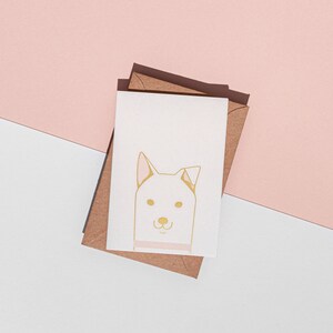 May include: A greeting card with a gold-colored dog illustration on a white background, resting on a brown envelope. The dog has pink accents on its ears and a pink collar. The background is split between white and light pink.