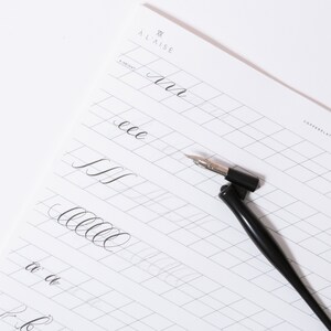 Pu&ograve; includere: Un foglio di pratica di calligrafia con il marchio "&Agrave; LAISE" e la parola "COPPERPLATE" stampati in alto a destra. Il foglio presenta un motivo a griglia ed esempi di forme di lettere corsive. Un portapenna nero con un pennino argentato &egrave; posizionato sul foglio.