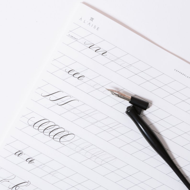Pu&ograve; includere: Un foglio di pratica di calligrafia con il marchio "&Agrave; LAISE" e la parola "COPPERPLATE" stampati in alto a destra. Il foglio presenta un motivo a griglia ed esempi di forme di lettere corsive. Un portapenna nero con un pennino argentato &egrave; posizionato sul foglio.