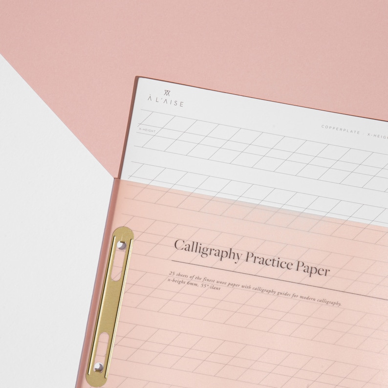 May include: Pink calligraphy practice paper with a gold-colored clasp. The paper has a grid pattern and the text "Calligraphy Practice Paper". The paper is inside a pink folder with the brand name "&Agrave; L'AISE" at the top.