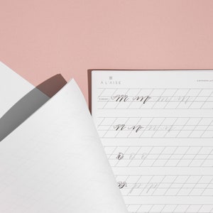 Pu&ograve; includere: Un blocco per esercitarsi nella calligrafia con il testo "&Agrave; L'AISE" e "COPPERPLATE" stampato in alto a destra. Il blocco &egrave; aperto su una pagina con la lettera "m" scritta in corsivo. Lo sfondo &egrave; rosa chiaro.