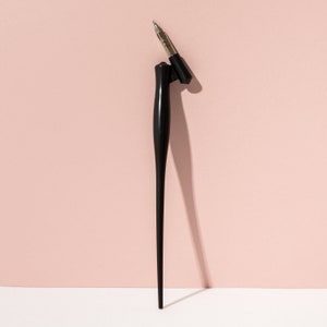Pu&ograve; includere: Un portapenna da calligrafia nero con pennino argentato e cappuccio nero. Il portapenna ha un design lungo e sottile, e il cappuccio &egrave; tolto, rivelando il pennino. Lo sfondo &egrave; rosa tenue e bianco.