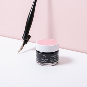 Pu&ograve; includere: Una penna stilografica nera con pennino argentato &egrave; posizionata vicino a un piccolo vasetto di inchiostro nero con coperchio rosa. Il vasetto riporta la scritta "Modern Calligraphy" e "15ml / 0,51 fl oz". Lo sfondo &egrave; rosa tenue.