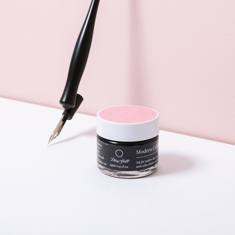 Pu&ograve; includere: Una penna stilografica nera con pennino argentato &egrave; posizionata vicino a un piccolo vasetto di inchiostro nero con coperchio rosa. Il vasetto riporta la scritta "Modern Calligraphy" e "15ml / 0,51 fl oz". Lo sfondo &egrave; rosa tenue.