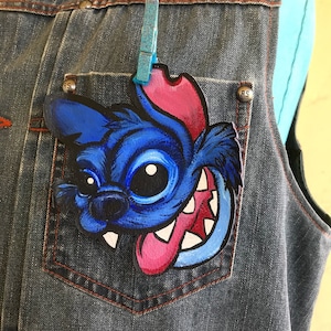 May include: A blue and pink cartoon character with a toothy grin is painted on a denim jacket. The character has a large, round eye and a long, pink tongue sticking out.
