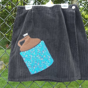 May include: A black corduroy skirt with a brown and blue applique of a jug. The jug has a blue and white patterned design.