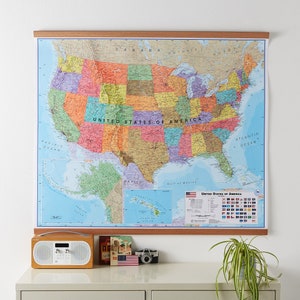 May include: A wall map of the United States of America with a wooden frame. The map shows the states in different colors and includes the names of the states, major cities, and bodies of water. The map also includes a smaller inset map of Alaska and Hawaii. The map is hanging on a white wall above a wooden table with a radio, a book, and a camera on it.