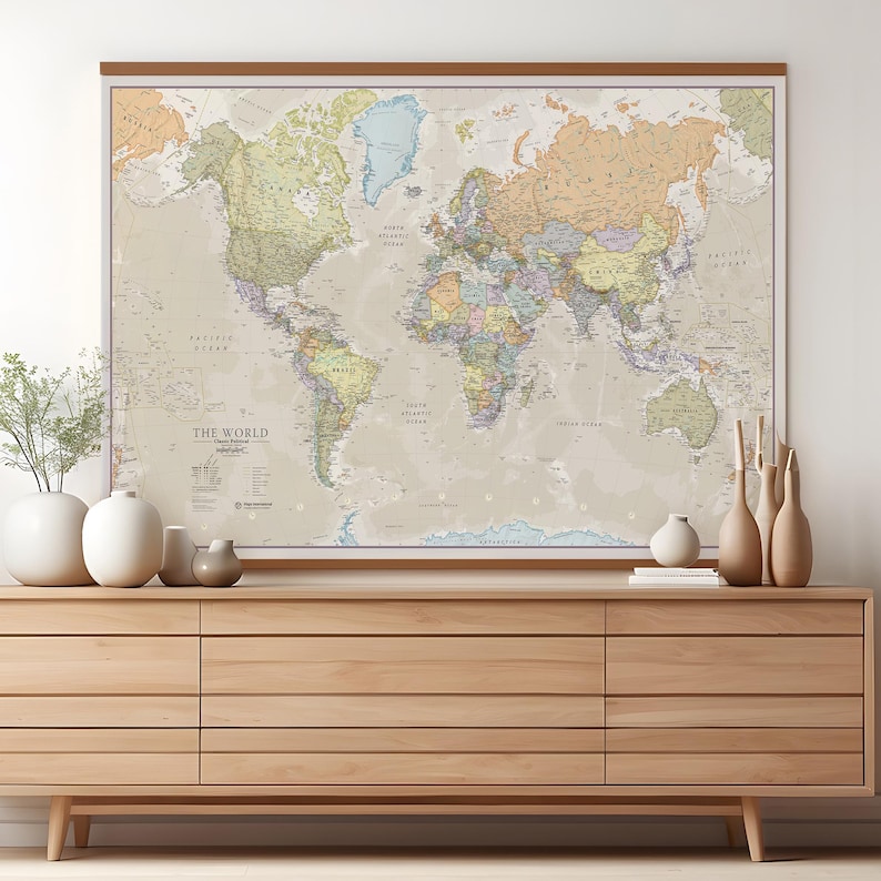 May include: A large, detailed world map with a beige background and colorful country borders. The map is framed in a light brown wood frame and is hanging on a wall above a light brown wooden dresser.