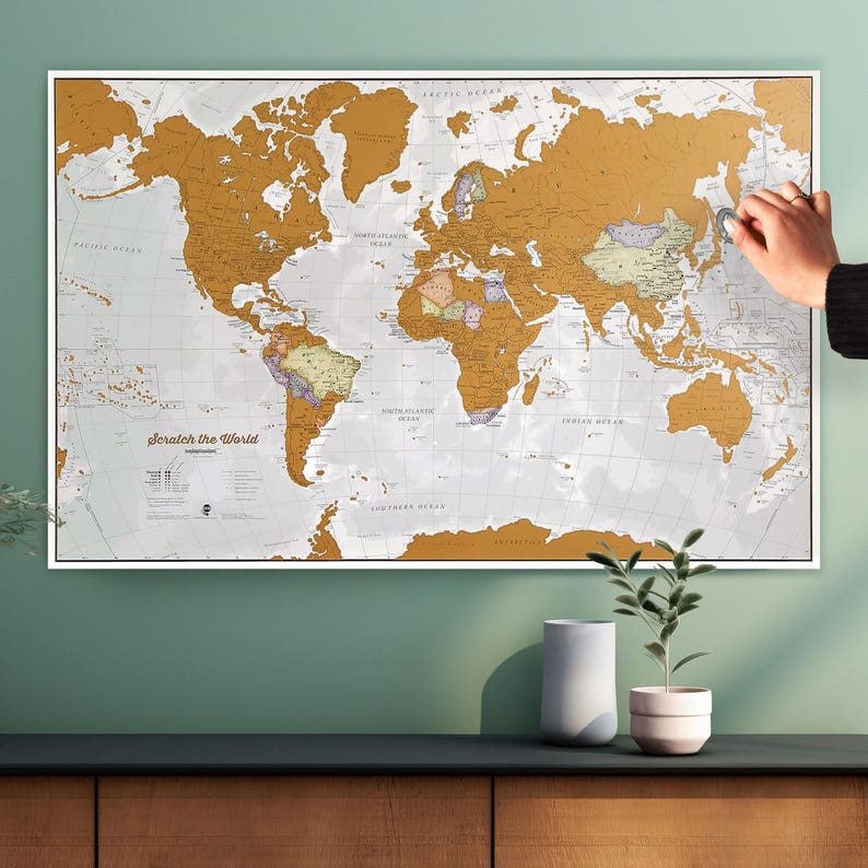 May include: A gold scratch-off world map with a white background. The map is mounted on a white frame and is hanging on a green wall. The map has the text "Scratch the World" printed on it.