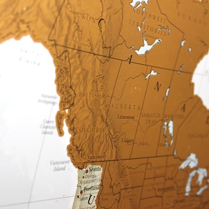 May include: A close-up of a gold-coloured map of North America, showing the provinces and states of Canada and the United States. The map highlights the Rocky Mountains, the Great Lakes, and the major cities of the region. The map is printed on a textured surface.