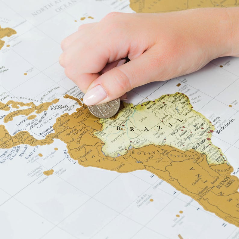 May include: A gold scratch-off world map with a hand holding a coin over South America. The map shows the continents and countries of the world. The gold colour is rubbed off in some areas, revealing the map underneath.