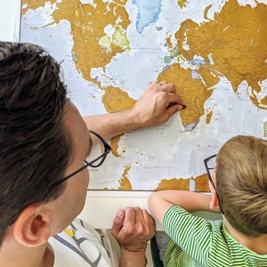 May include: A father and son are looking at a gold foil world map. The father is pointing at Africa with his right hand. The son is wearing glasses and looking at the map.