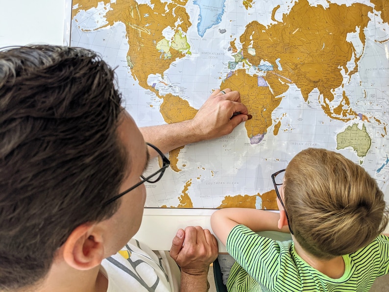 May include: A father and son are looking at a gold foil world map. The father is pointing at Africa with his right hand. The son is wearing glasses and looking at the map.