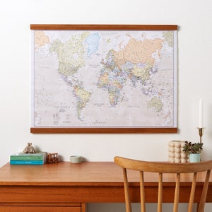May include: A world map with a wooden frame hangs on a white wall above a wooden desk with a chair. The map is beige with colorful countries and oceans. The desk has a small wooden toy car, a small pyramid, and three books on it. There is a small potted plant and a ceramic vase on the desk.