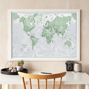 May include: A framed world map with a white background and green landmasses. The map is hanging on a light wood wall above a white desk with a wooden chair, a camera, and a small plant.