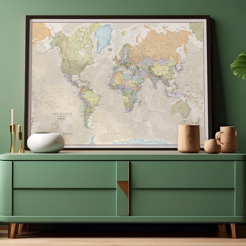 May include: A framed world map with a beige background and colorful country borders. The map is hanging on a green wall above a green dresser with two drawers. There are three gold candle holders and a white ceramic bowl on the dresser. Two wooden mugs are on the dresser to the right of the map.