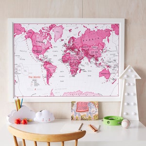 May include: A pink world map in a white frame hangs above a white desk with a white cloud-shaped pencil holder, a white cloud-shaped pencil holder with a smiley face, a book with the text "E is for Elephant", a green tambourine, and a baseball. The map has the text "The World" in the lower left corner.