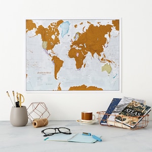 May include: A gold scratch-off world map with a white background. The map is framed and hanging on a white wall. There is a copper wire basket with books and a coffee cup on a white table in the foreground.