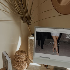 May include: A computer screen displaying a website for Abilene Photography, featuring a couple walking down a street. The website has a white background with black text. The computer is on a white desk with a book and a woven basket.