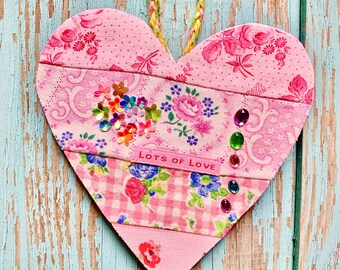 May include: A pink heart-shaped wall hanging with a floral fabric design and the words "LOTS OF LOVE". The heart is decorated with sequins and gemstones, and has a braided rope for hanging.