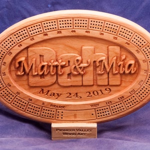 May include: A wooden oval cribbage board with a carved design of the names "Matt & Mia" and the date "May 24, 2019". The board has a traditional cribbage board layout with numbered holes and a scoring area.