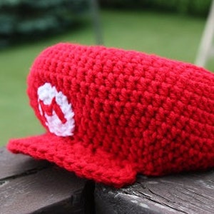 May include: Red crocheted hat with a white 'M' on the front. The hat has a brim and is made with a textured stitch.