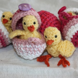 May include: Four crocheted chicks, two in pink and white striped egg-shaped baskets, one in a pink egg-shaped basket, and one standing on its own. All chicks are yellow with orange beaks and black eyes.