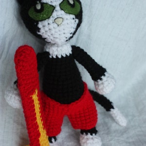 May include: A black and white crocheted cat wearing red shorts and holding a yellow and red surfboard.