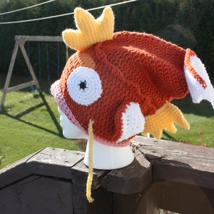 May include: A crocheted hat shaped like a red and orange fish with white and yellow details. The hat is sitting on a wooden surface.