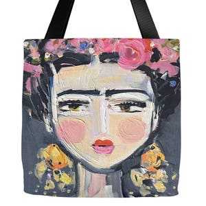 May include: A tote bag featuring a colorful painting of a woman with a flower crown. The woman has dark hair, brown eyes, and pink cheeks. The background is a dark gray.