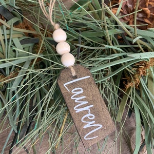 May include: A wooden Christmas ornament with the name "Lauren" written in white paint. The ornament is decorated with a string of white wooden beads and twine.
