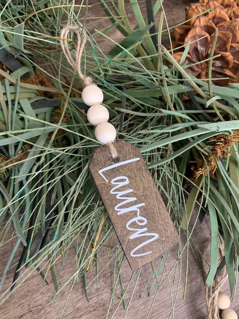 May include: A wooden Christmas ornament with the name "Lauren" written in white paint. The ornament is decorated with a string of white wooden beads and twine.