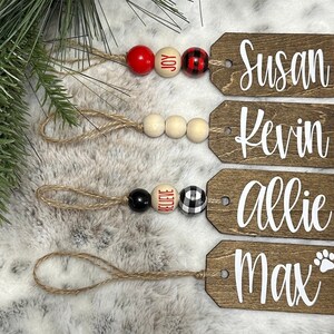 May include: Four wooden gift tags with twine and beads. The tags are decorated with the names "Susan", "Kevin", "Allie", and "Max" with a paw print. The beads are red, white, black, and buffalo plaid.