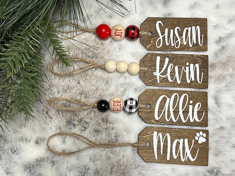 May include: Four wooden gift tags with twine and beads. The tags are decorated with the names "Susan", "Kevin", "Allie", and "Max" with a paw print. The beads are red, white, black, and buffalo plaid.