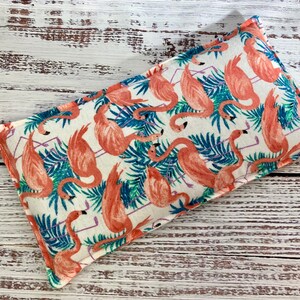 May include: A white fabric rectangular pillow with a pink flamingo print. The pillow has a white background with pink flamingos and blue palm leaves.