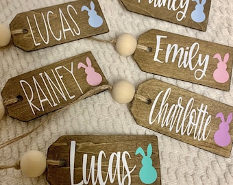 May include: Five wooden Easter tags with bunny silhouettes and names written in white. The names are Lucas, Rainey, Emily, Charlotte, and Lucas.
