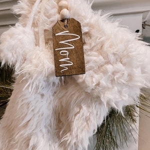 May include: White faux fur stocking with a wooden tag that says "Mom" and a string of wooden beads.
