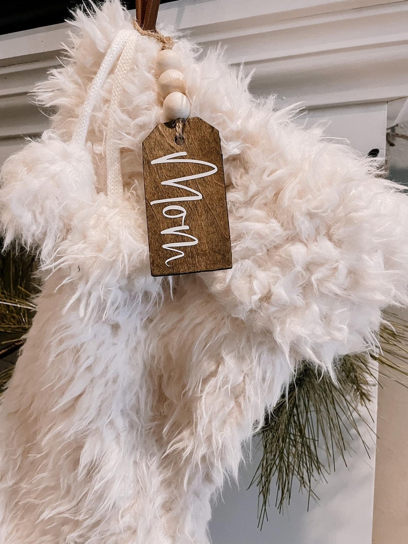 May include: White faux fur stocking with a wooden tag that says "Mom" and a string of wooden beads.