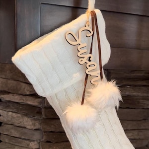 May include: A white cable-knit Christmas stocking with a wooden name tag that says "Susan". The tag is attached with brown leather straps and fluffy white pom-poms.