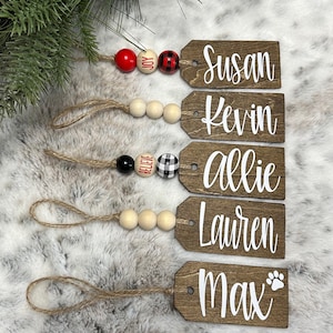 May include: Five wooden Christmas ornaments with names written in white on them. The ornaments are strung together with twine and wooden beads. The beads are red, white, black, and buffalo plaid.