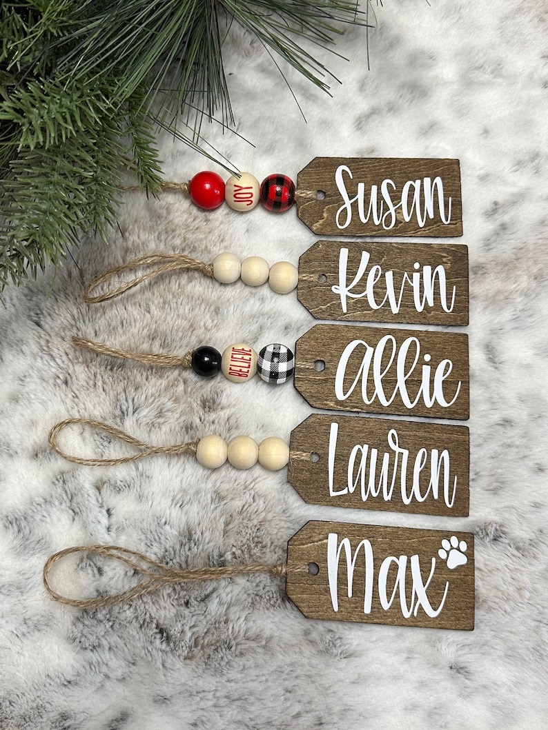 May include: Five wooden Christmas ornaments with names written in white on them. The ornaments are strung together with twine and wooden beads. The beads are red, white, black, and buffalo plaid.
