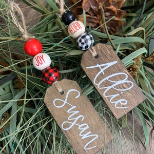 May include: Two wooden Christmas ornaments with names "Susan" and "Allie" written in white paint. Each ornament is decorated with a string of beads, including red, white, black, and buffalo plaid.