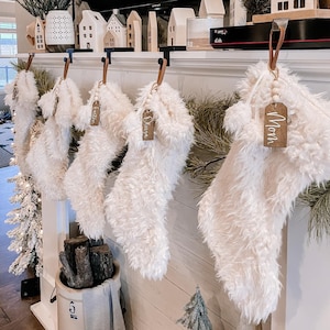 May include: Five white fuzzy Christmas stockings with wooden tags that have names written on them. The names are Olivia, Logan, Noah, and two others that are not visible. The stockings are hanging on a white mantel with a wooden background.